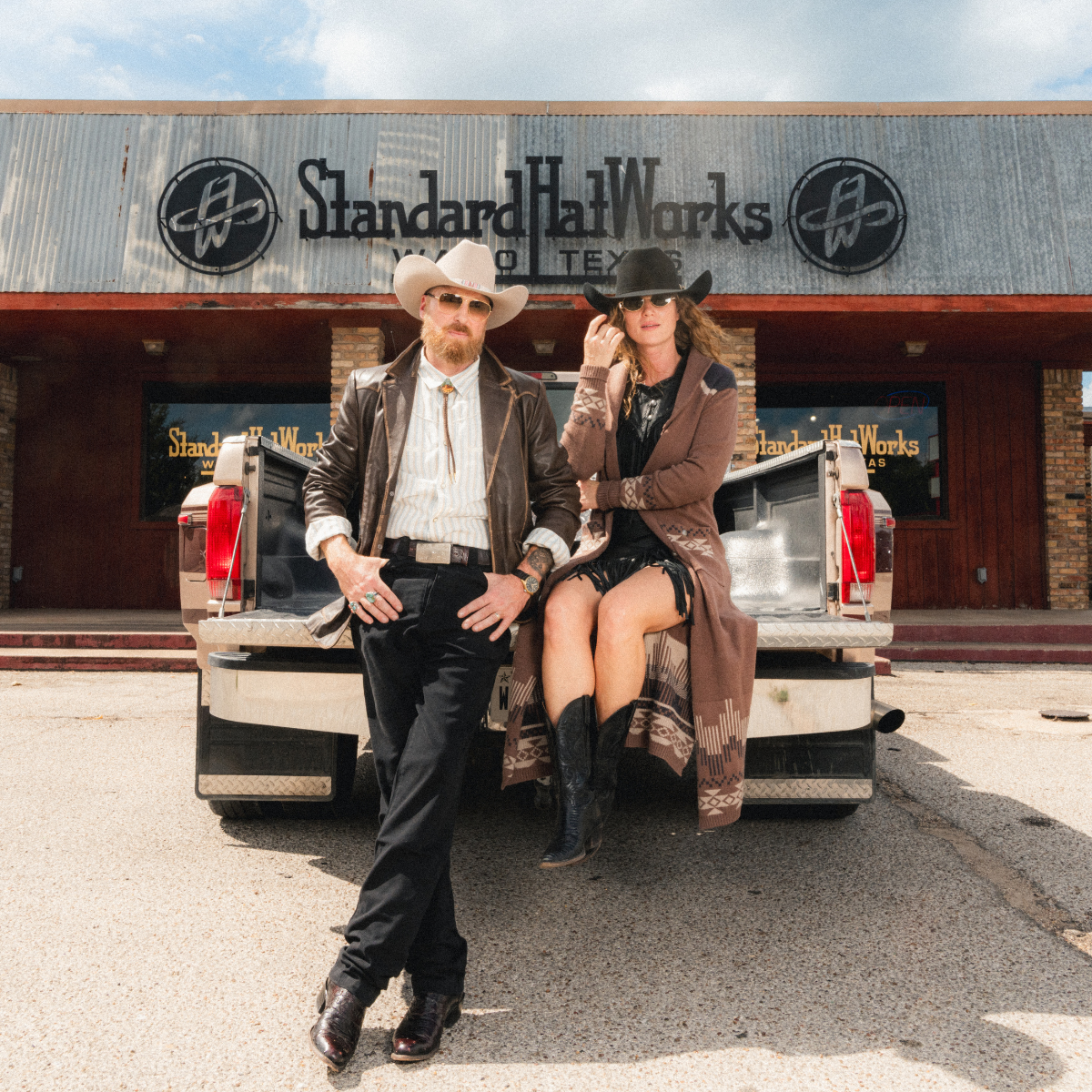 A man and a woman in western-style clothing pose in front of a pickup truck parked outside the 'Standard Hat Works' building. The man wears a roper felt cowboy hat, our David Allen leather jacket, the Cash pearl snap shirt, and bolo tie, while the woman wears a black cattleman felt hat, winona knitted duster cardigan, and black cowboy boots.